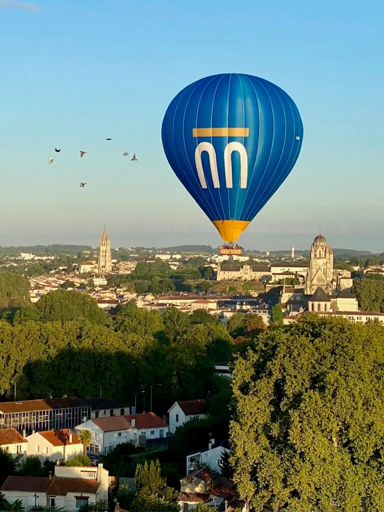 Une montgolfière bleue à rayures blanches verticales. L'inscription « Le Bonheur c'est si SAINTES » est imprimée en blanc sur le ballon.