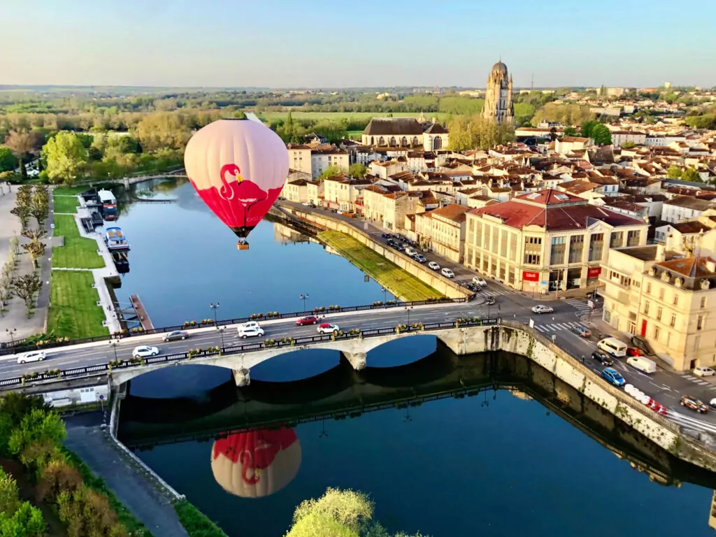 Une montgolfière survole une rivière avec un pont de pierre à trois arches dans une ville pittoresque.